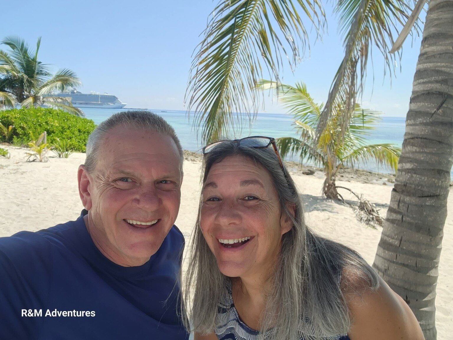 Ron and Michelle smiling beneath palm trees at Ocean Cay with a cruise ship behind them